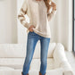 A person wears a Shewin Khaki Cable Knit Colorblock Crew Neck Drop Shoulder Sweater, jeans, and brown boots, standing indoors by a white couch and smiling with one hand in their pocket.