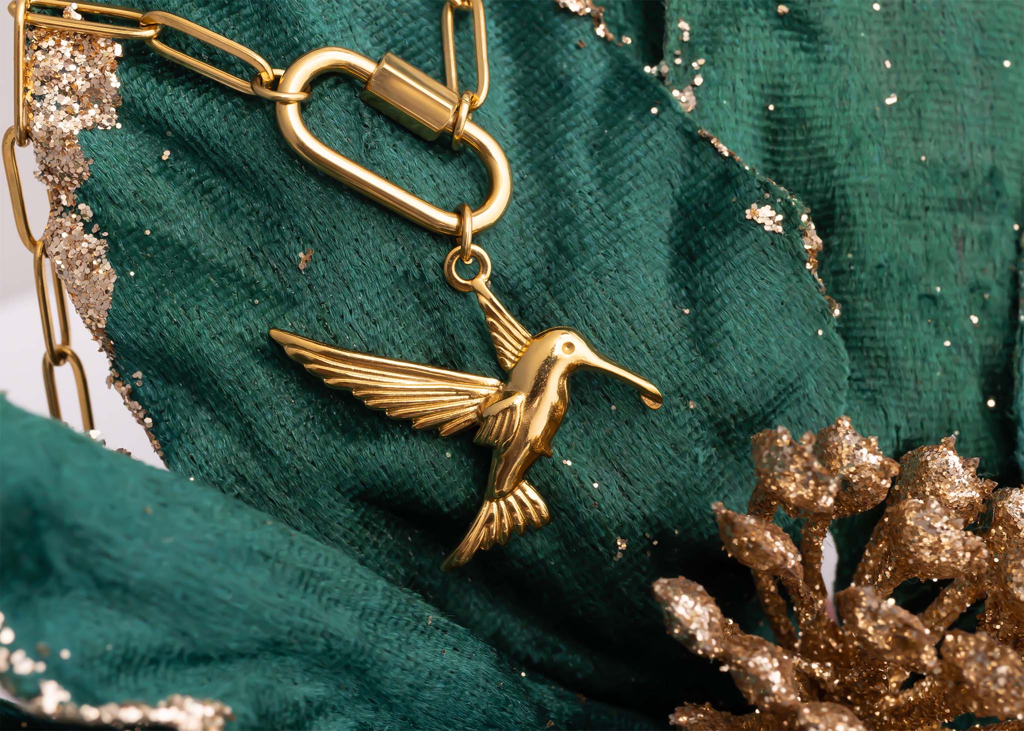 WJW 18K Gold PVD Paperclip Carabiner Necklace featuring a gold hummingbird pendant, displayed on green textured fabric with gold glitter accents and a decorative gold pinecone.