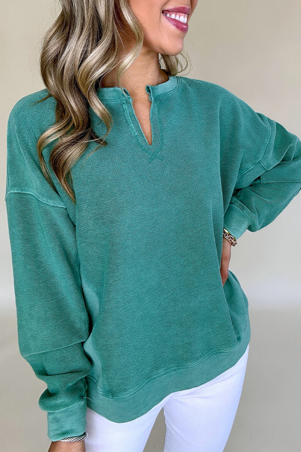 A person wearing the comfortable Shewin Notched Neck Exposed Seam Drop Shoulder Sweatshirt in green, styled with long sleeves, stands with one hand on their hip, paired with white pants.