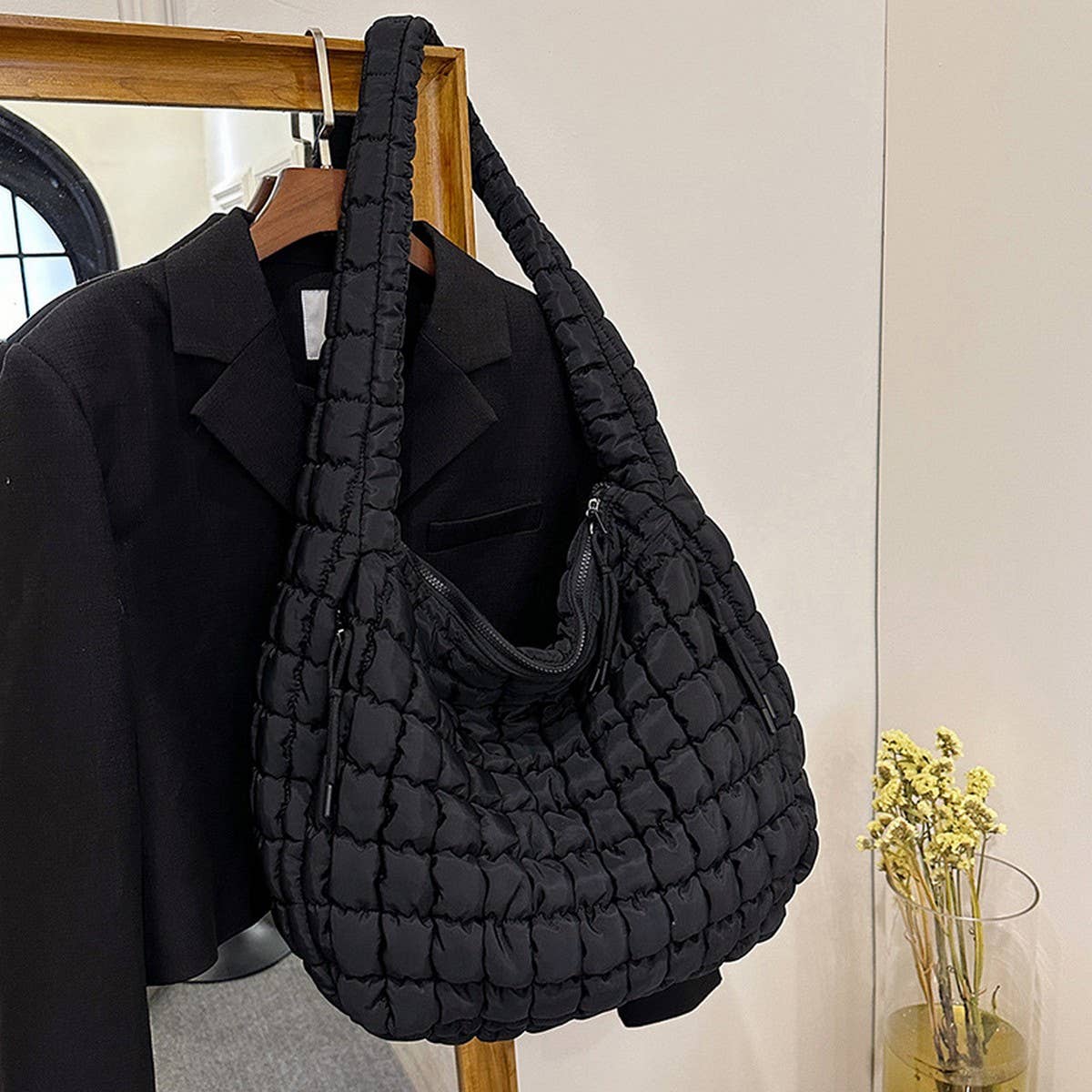 The ACCITY Cloud Tote Bag Large Shoulder Crossbody in black with a quilted design hangs on a wooden rack beside a black blazer; nearby, yellow dried flowers rest on a table.