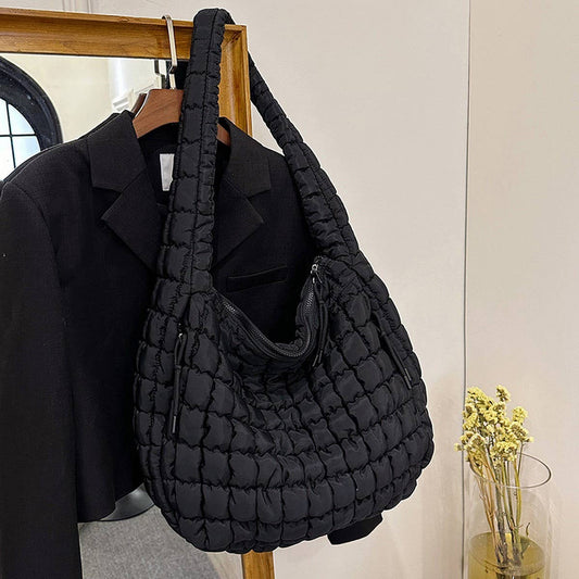 The ACCITY Cloud Tote Bag Large Shoulder Crossbody in black with a quilted design hangs on a wooden rack beside a black blazer; nearby, yellow dried flowers rest on a table.