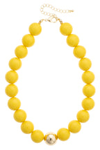 The Marjorie Game Day Beaded Necklace by Canvas Style is a bold statement piece featuring large, round yellow oversized beads and a single metallic gold bead at the center. It is finished with an adjustable gold chain clasp, making it perfect for adding a pop of color to any outfit.