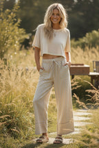 A woman stands outdoors on a stone path wearing Sewn and Seen's Woven Striped Wide Leg Pants, a beige short-sleeve top, and sandals. Tall grass and coastal-inspired outdoor furniture appear in the background.