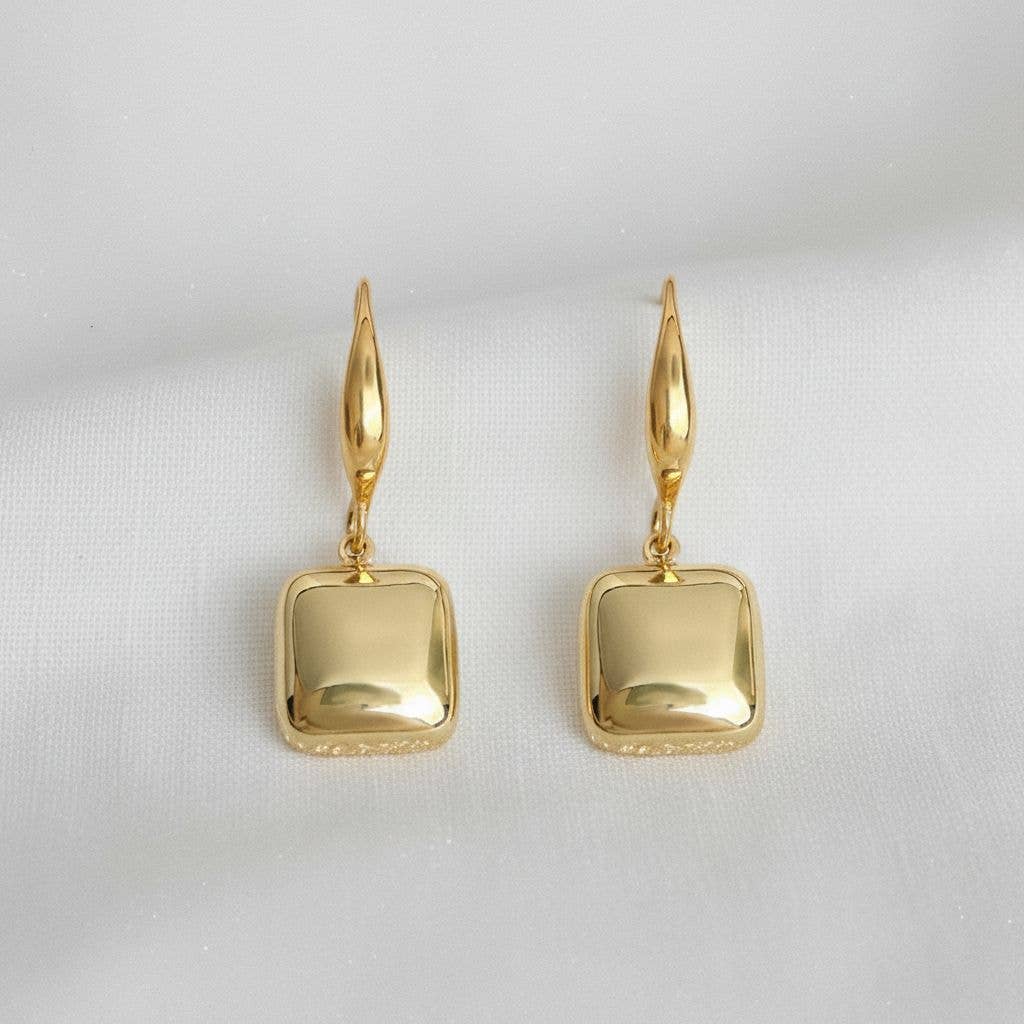 A pair of Ellemadera Modern Geometric Drop Earrings with rounded square-shaped gold pendants displayed on a white fabric background.