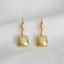A pair of Ellemadera Modern Geometric Drop Earrings with rounded square-shaped gold pendants displayed on a white fabric background.
