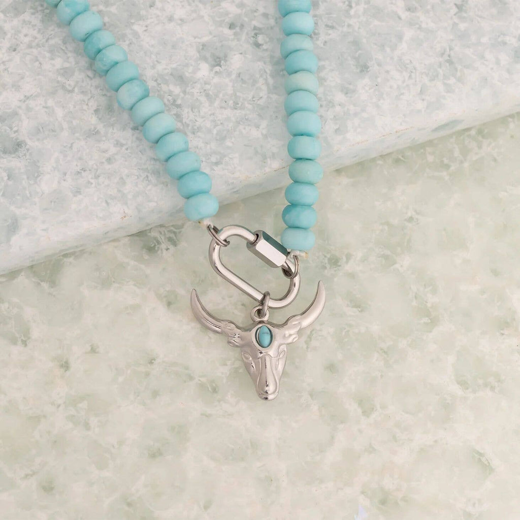 The WJW Stainless Steel Turquoise Chunky Bead Carabiner Necklace features vibrant turquoise beads and a striking silver bull skull pendant accented with a small turquoise stone. Designed for durability, this unique necklace showcases a secure carabiner clasp closure and is beautifully displayed on a light textured surface.