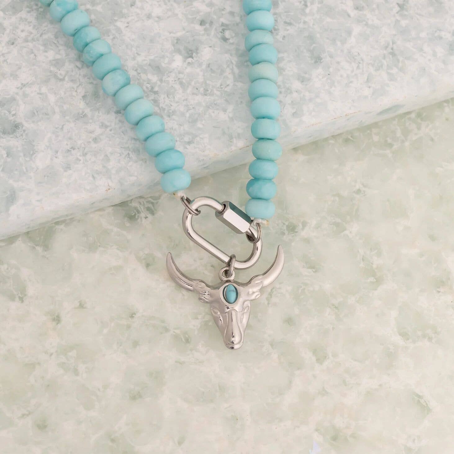 The WJW Stainless Steel Turquoise Chunky Bead Carabiner Necklace features vibrant turquoise beads and a striking silver bull skull pendant accented with a small turquoise stone. Designed for durability, this unique necklace showcases a secure carabiner clasp closure and is beautifully displayed on a light textured surface.
