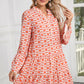 A person stands indoors wearing the Orange Floral Button V Neck Bell Sleeve Mini Dress by Shewin, showcasing long wavy hair and floral earrings.