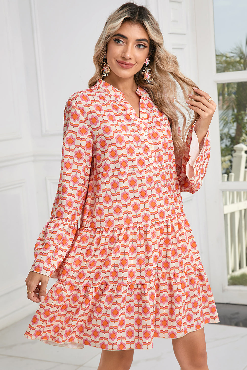 A person stands indoors wearing the Orange Floral Button V Neck Bell Sleeve Mini Dress by Shewin, showcasing long wavy hair and floral earrings.