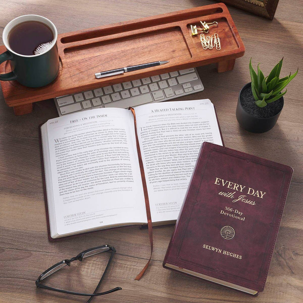 An open Devotional Every Day with Jesus Faux Leather next to a laptop, a mug of coffee, a small succulent, and a tray of paperclips on a wooden desk.