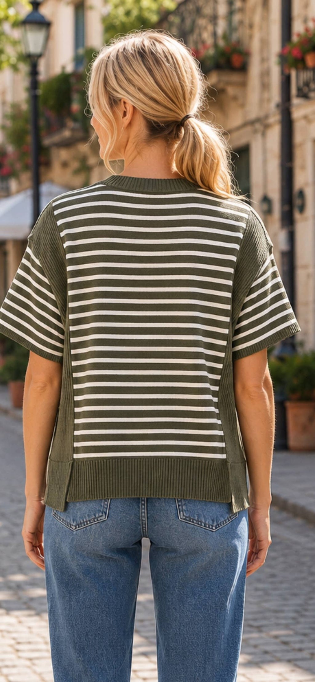 A person with blonde hair in a ponytail is standing outdoors on a cobblestone street, wearing blue jeans and the Celeste Clothing Women’s Stripe Color Block Rib Knit Relaxed Top—a green and white striped short-sleeve casual summer top made from rib knit fabric.