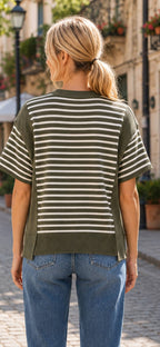 A person with blonde hair in a ponytail is standing outdoors on a cobblestone street, wearing blue jeans and the Celeste Clothing Women’s Stripe Color Block Rib Knit Relaxed Top—a green and white striped short-sleeve casual summer top made from rib knit fabric.