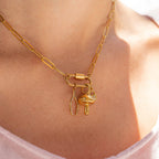 Close-up of the WJW 18K Gold PVD Paperclip Carabiner Necklace, featuring three charms—a crystal-encrusted leaf, a gold mushroom, and a gold capsule—worn by a person in a light pink top.