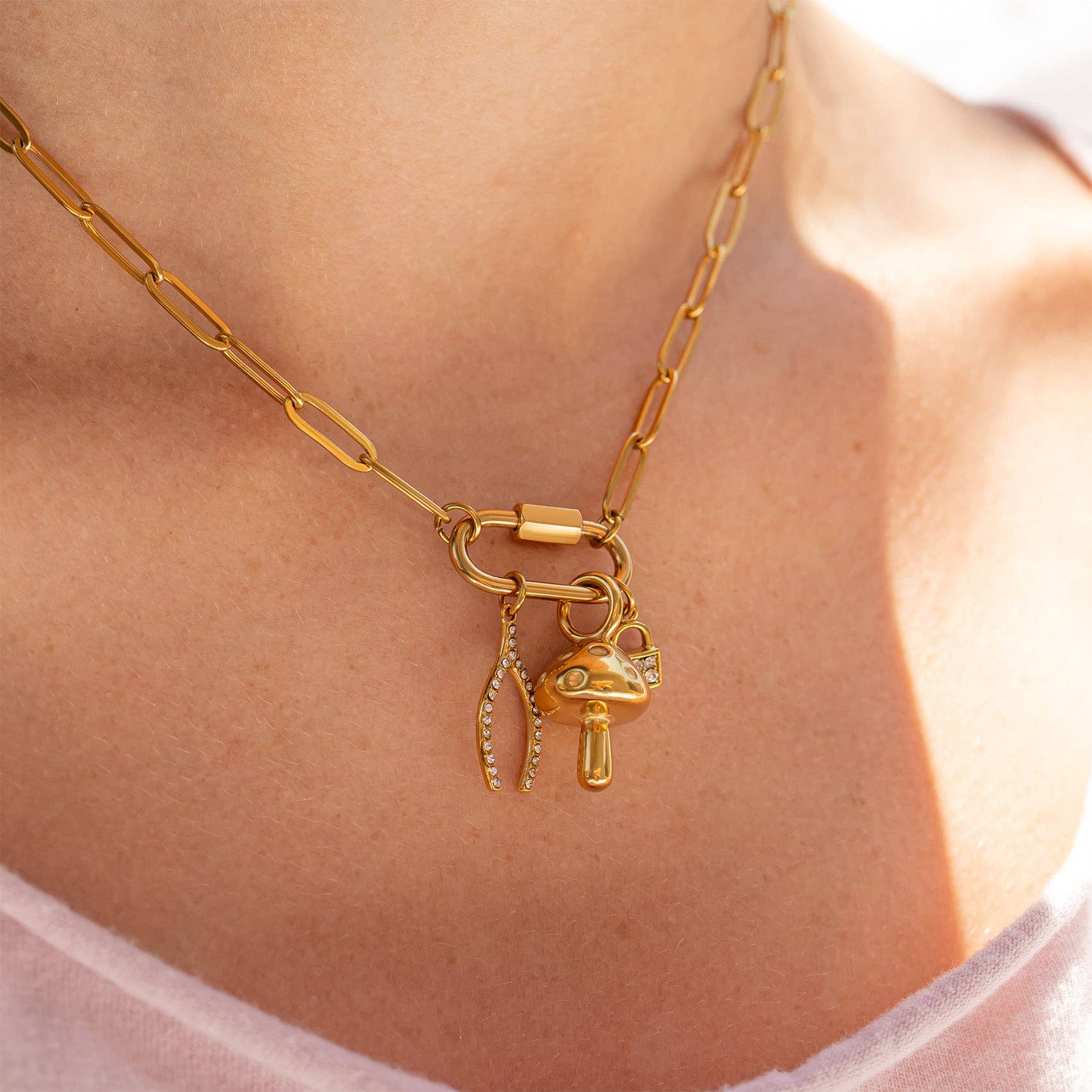 Close-up of the WJW 18K Gold PVD Paperclip Carabiner Necklace, featuring three charms—a crystal-encrusted leaf, a gold mushroom, and a gold capsule—worn by a person in a light pink top.