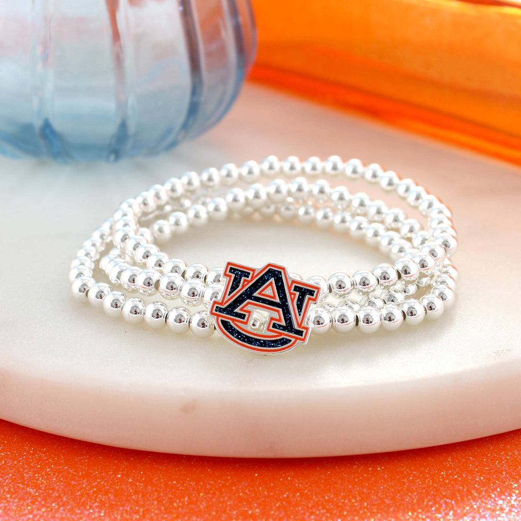 The Auburn Glitter Enamel Logo Stretch Bracelet Set by Seasons Jewelry showcases an officially licensed silver beaded design with the Auburn University logo charm accented in glitter enamel, displayed on a white round surface beside a blue glass object and orange background elements.