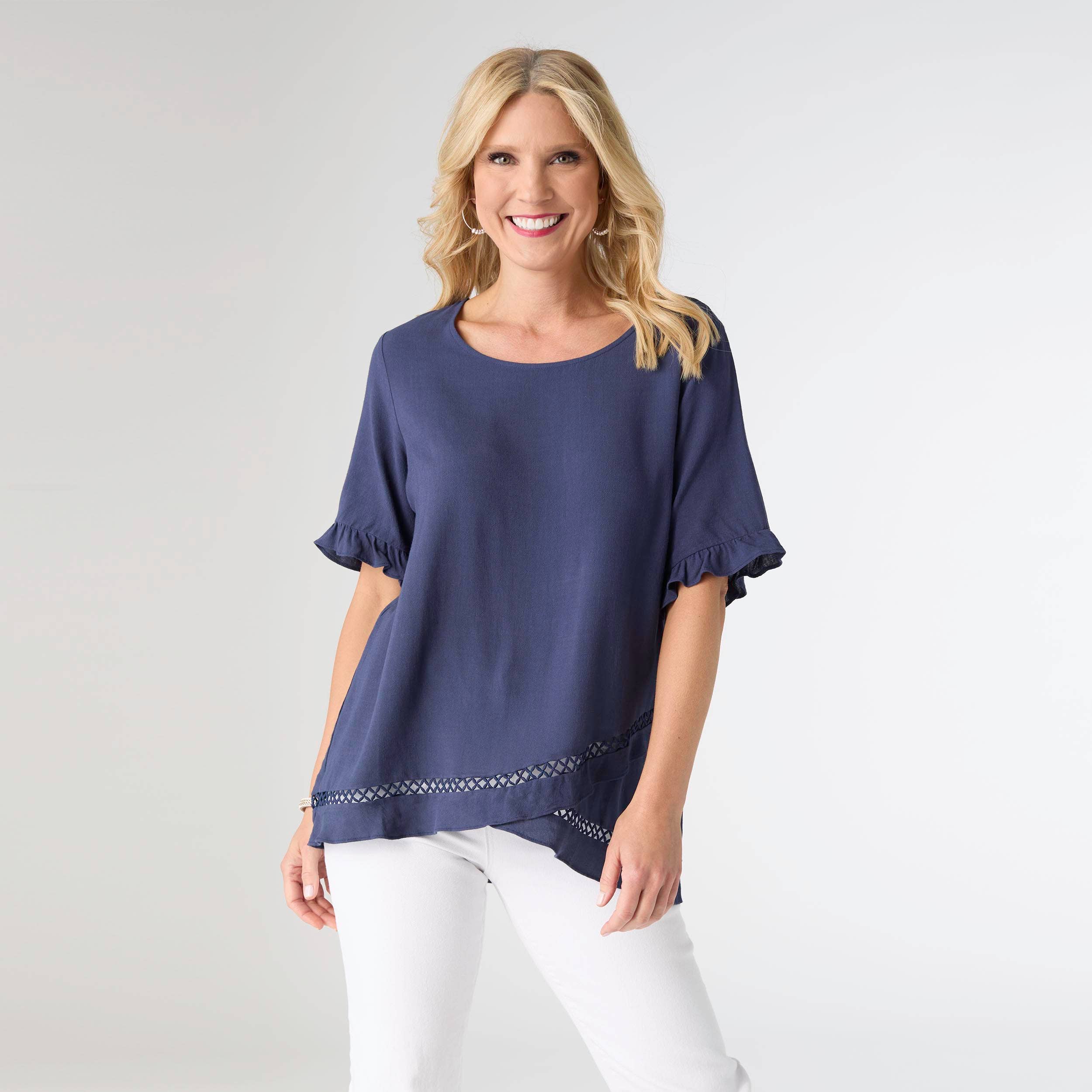 A woman with blonde hair smiles in the COCO + CARMEN Layla Short Sleeve Crossover Ruffle Top (navy blue) and white pants, standing against a plain light background. Main image