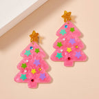 Add a touch of festive sparkle with the Glittery Christmas Tree Drop Earrings by Lilla Haven. These holiday statement earrings, crafted from pink glitter acrylic and adorned with colorful star and jewel decorations topped with a shimmering gold star, are the perfect accessory for your celebrations. The Lilla Haven Holiday Acrylic Earrings are displayed here on a beige surface.