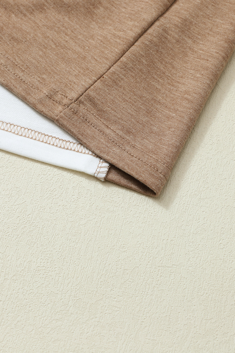 Close-up of the hem of the Shewin Khaki Color Block Exposed Seam Long Sleeve Top, showcasing the stitching detail and a white inner layer. The fabric, known for its versatile khaki color, lies flat on a light-colored surface.