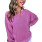 A woman smiles while wearing a Notched Neck Exposed Seam Drop Shoulder Sweatshirt by Shewin in pink, paired with white pants, standing against a plain white background.