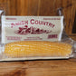 A packaged corn cob labeled "White Popcorn Microwave Cobs" from Amish Country Popcorn, ideal for popping fresh popcorn, sits on a wooden surface.