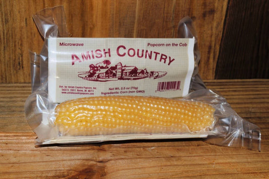A packaged corn cob labeled "White Popcorn Microwave Cobs" from Amish Country Popcorn, ideal for popping fresh popcorn, sits on a wooden surface.