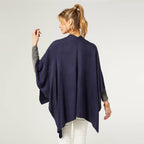 A woman faces away, wearing the COCO + CARMEN Kiara Lightweight Wrap in navy blue over a gray long-sleeve shirt and white pants, set against a plain light background.