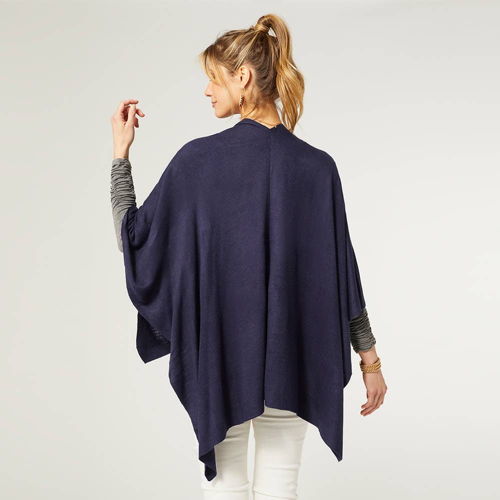 A woman faces away, wearing the COCO + CARMEN Kiara Lightweight Wrap in navy blue over a gray long-sleeve shirt and white pants, set against a plain light background.
