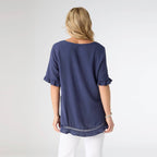 A woman faces away from the camera, wearing the COCO + CARMEN Layla Short Sleeve Crossover Ruffle Top in navy blue with white pants, set against a plain light background.