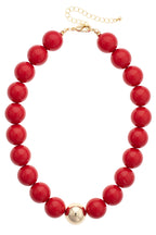 The Marjorie Game Day Beaded Necklace by Canvas Style features large, glossy red beads highlighted by a central gold bead, finished with a gold clasp and adjustable chain—an eye-catching statement piece perfect for adding bold flair to any outfit.