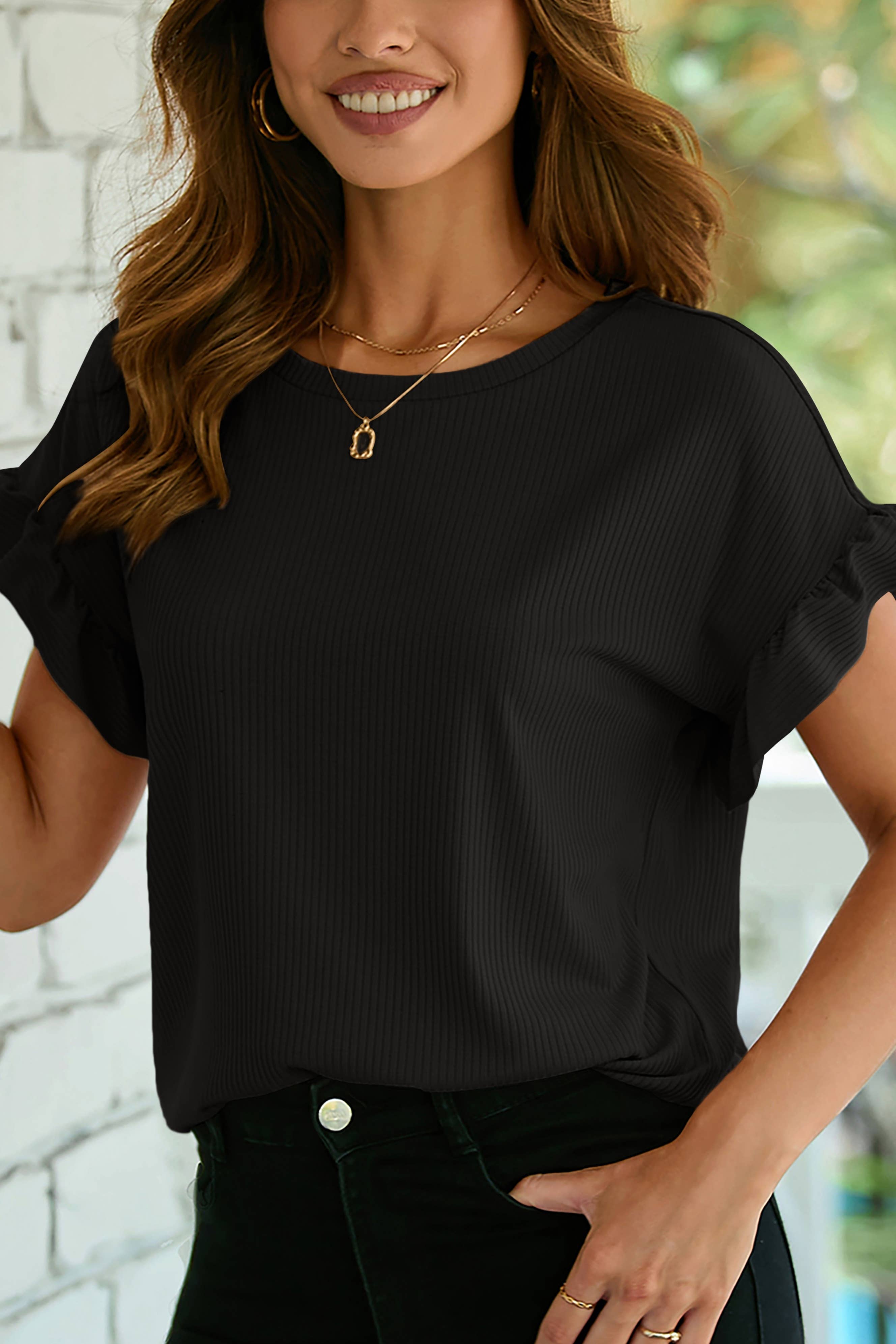 A woman wearing the 1 the Woman Solid Color Ribbed Ruffled Short Sleeve T Shirt with black pants and gold jewelry stands indoors by a white brick wall. Main image