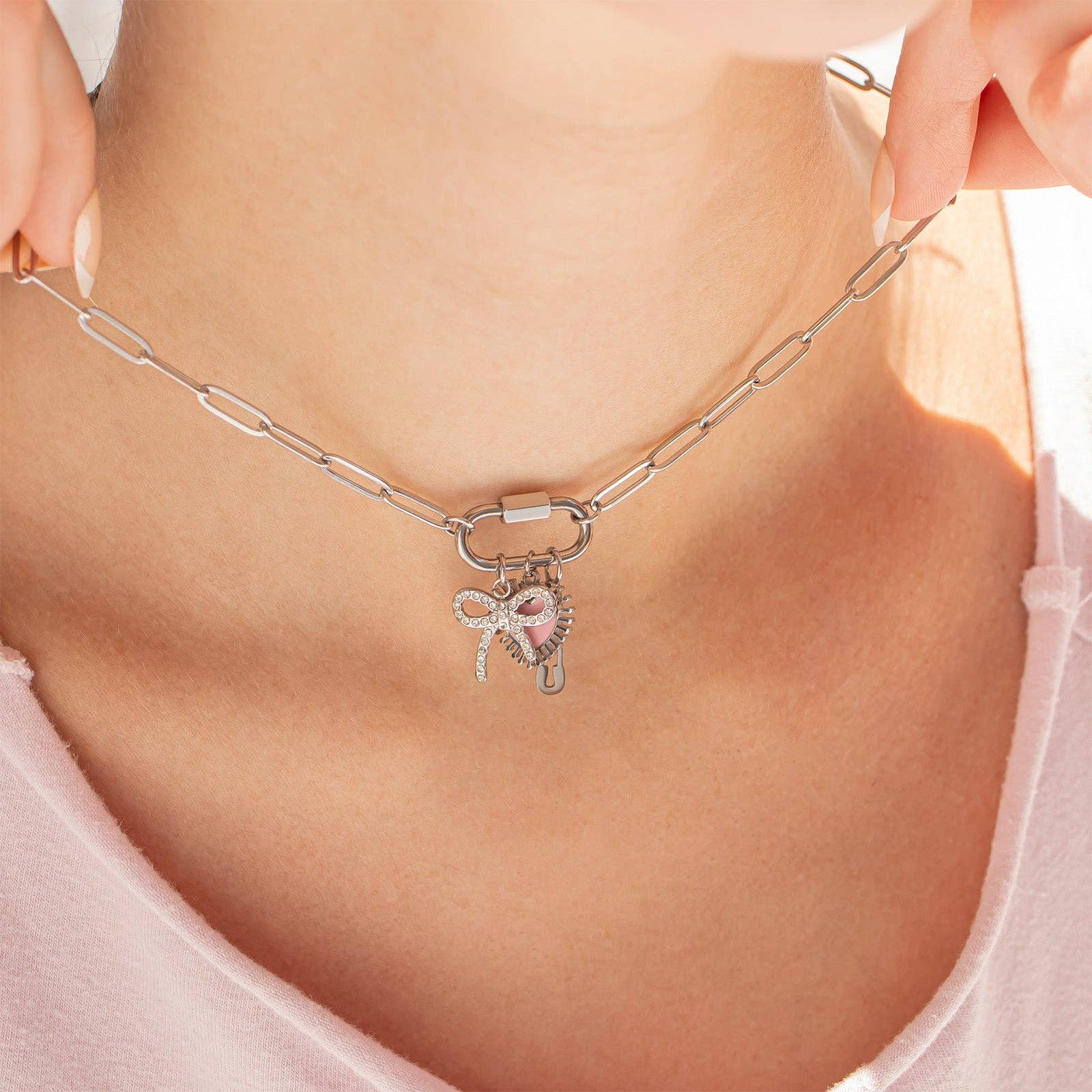 A person wearing the WJW 18K Gold PVD Paperclip Carabiner Necklace, featuring a heart-shaped pendant, key, and small bow charm, shown against light skin and a pink top.