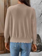 A person stands indoors facing away, wearing the Suhkasana Women’s Solid Color V-Neck Lettuce Edge Knit Cardigan in beige and blue jeans.