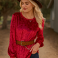A woman stands outdoors with one hand in her pocket, wearing the 24-Hrs Fashion Crushed Velvet Top in red with flowy sleeves, a brown hat and belt, and dark jeans.