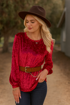 A woman stands outdoors with one hand in her pocket, wearing the 24-Hrs Fashion Crushed Velvet Top in red with flowy sleeves, a brown hat and belt, and dark jeans.