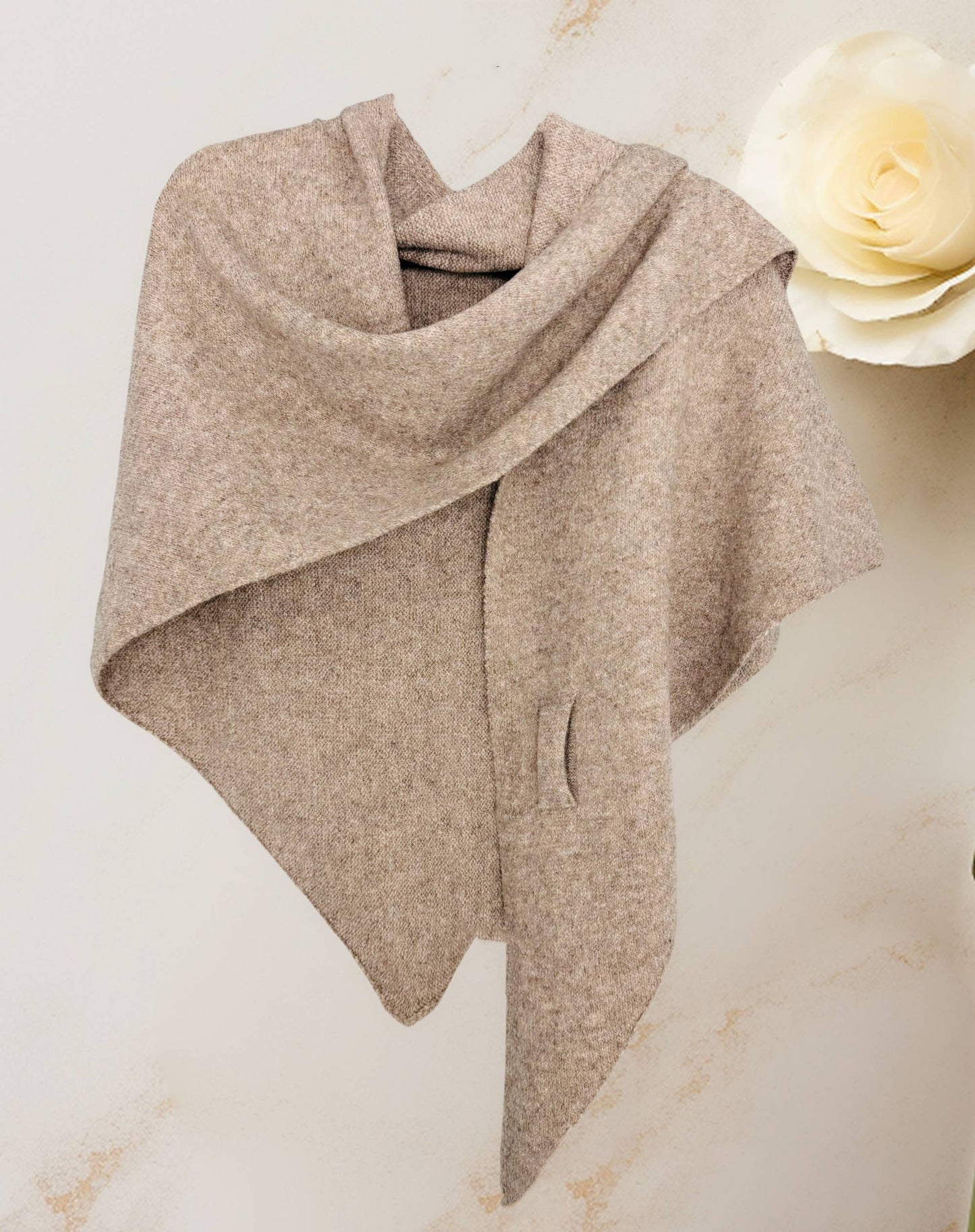 The JC Sunny Fashion Cashmere-Blend Knit Triangle Shawl with Cross-Front Design, in beige, is draped on a wall like a cozy scarf. A large ivory rose is seen in the background on the right.