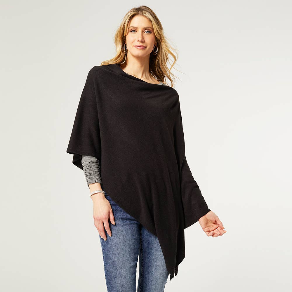 A woman models the COCO + CARMEN Lightweight Ponchos Wrap in black over a gray long-sleeve shirt and blue jeans, standing against a light background for a chic layered look.