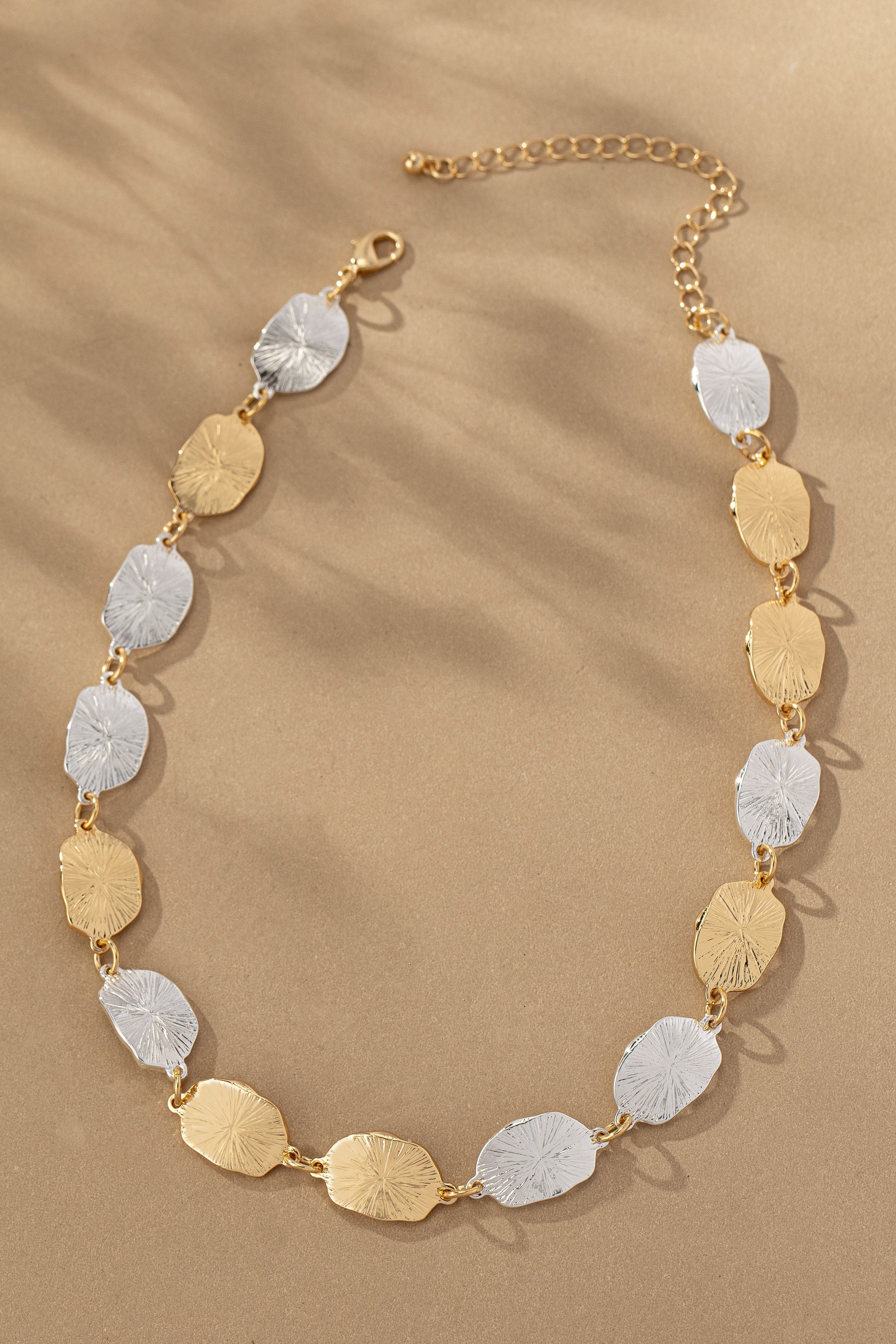 Two Tone Irregular Shape Hammered Beaded Necklace from FASHION GO arranged in a semi-circle with a solar ray pattern on a sandy background.