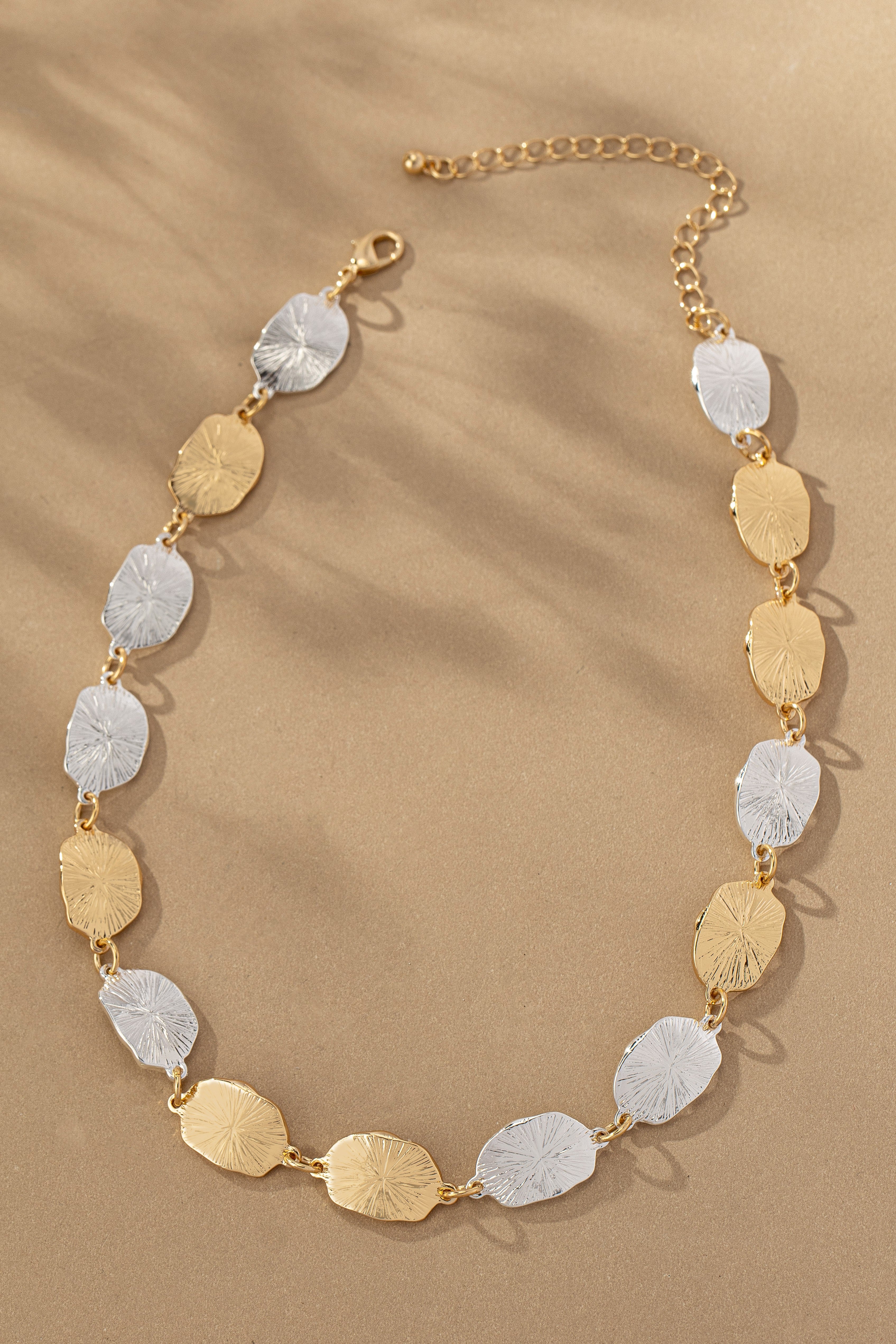 Two Tone Irregular Shape Hammered Beaded Necklace from FASHION GO arranged in a semi-circle with a solar ray pattern on a sandy background.