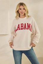 A person wearing a cream-colored, long-sleeved ALABAMA Cloudy Soft Knit Long Sleeve Top with “ALABAMA” printed in red across the chest.