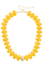 The San Sebastián Beaded Resin Necklace by Canvas Style features large round yellow resin beads accented with a single metallic centerpiece and finished with a gold adjustable chain clasp. This bold statement necklace is perfect for elevating any look.