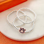 Three silver beaded bracelets from the Seasons Jewelry Auburn Glitter Enamel Logo Stretch Bracelet Set are displayed on a round white marble surface, with one bracelet featuring an officially licensed Auburn University “AU” logo charm accented with glitter enamel.