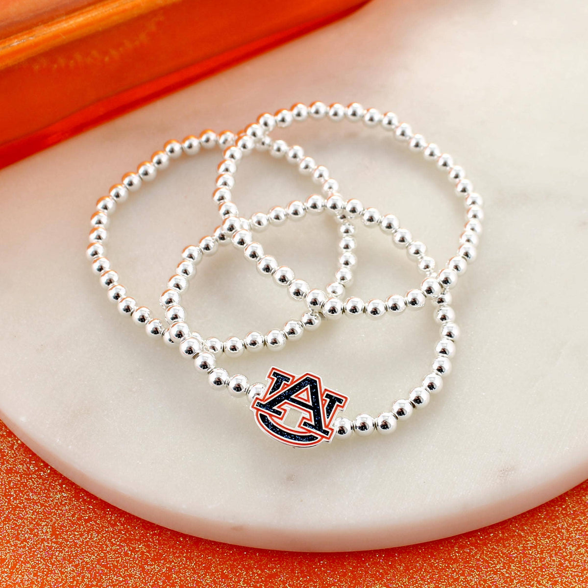 Three silver beaded bracelets from the Seasons Jewelry Auburn Glitter Enamel Logo Stretch Bracelet Set are displayed on a round white marble surface, with one bracelet featuring an officially licensed Auburn University “AU” logo charm accented with glitter enamel.