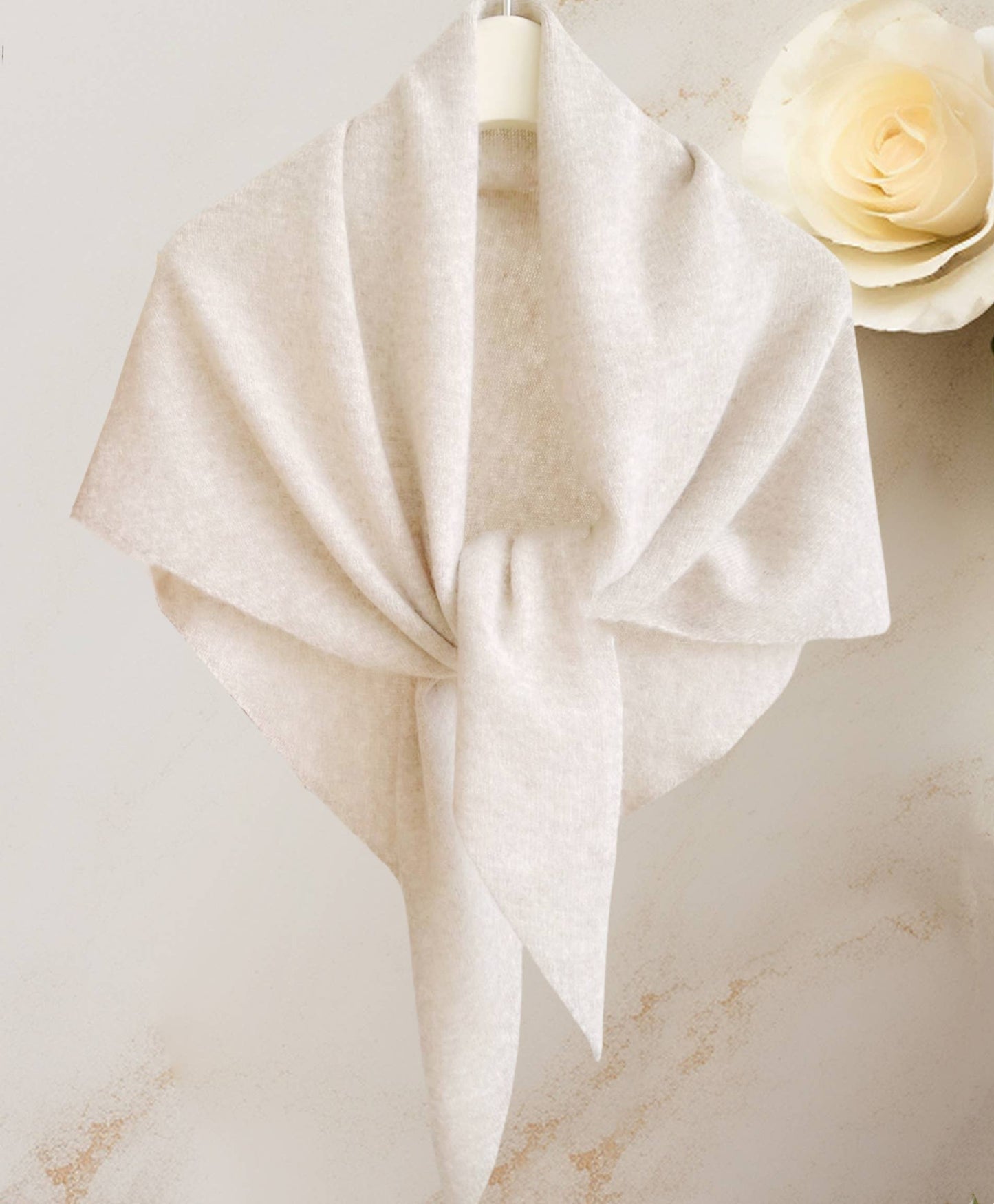 The JC Sunny Fashion Cashmere-Blend Knit Triangle Shawl with Cross-Front Design, in light beige, is displayed on a hanger tied loosely, with a cream rose in the background for chic style.