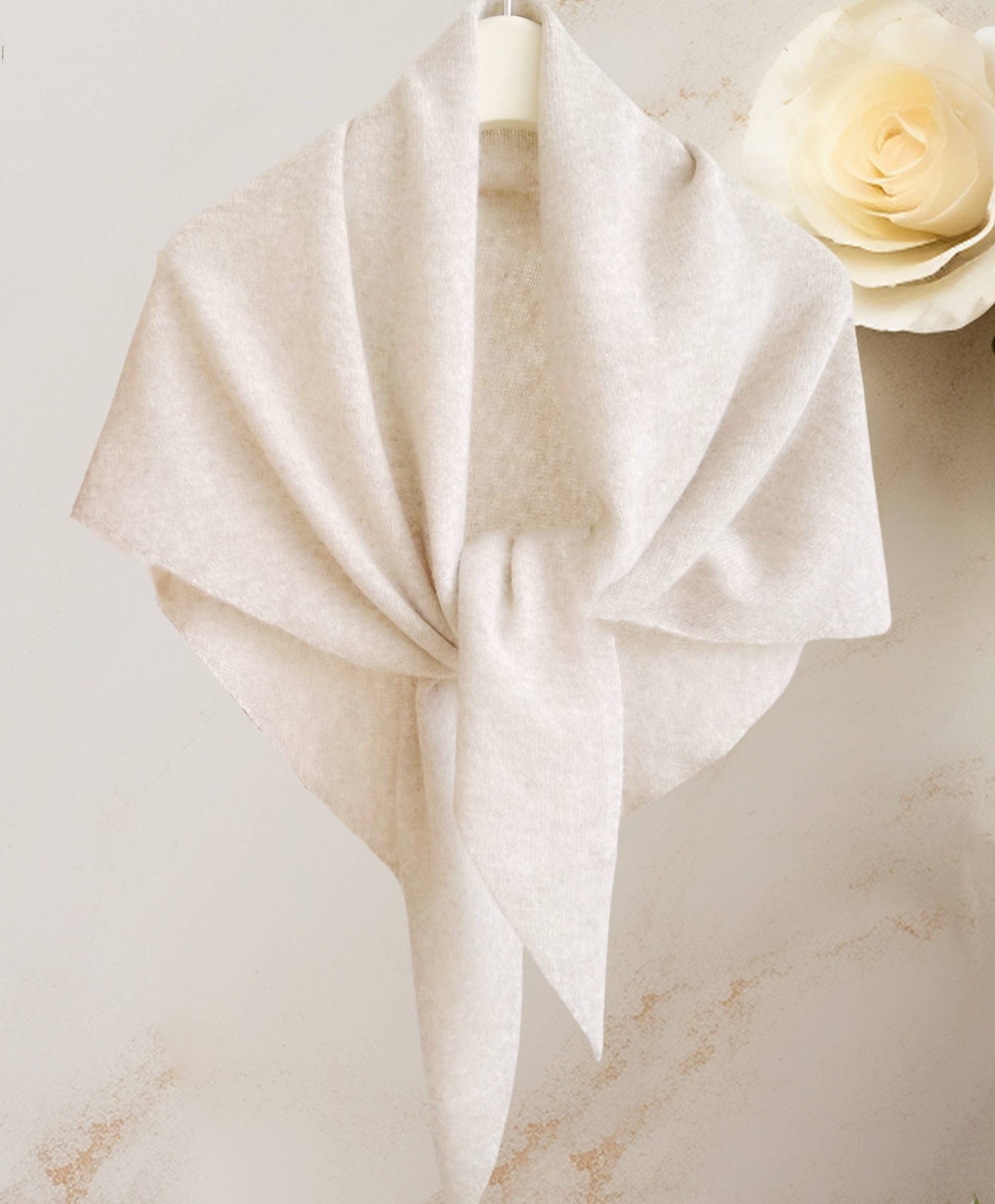 The JC Sunny Fashion Cashmere-Blend Knit Triangle Shawl with Cross-Front Design, in light beige, is displayed on a hanger tied loosely, with a cream rose in the background for chic style.