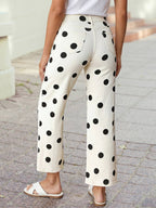 A person wearing 1 the Woman Polka Dot Print Straight Leg Casual Pants in white with black dots, paired with a white top and sandals, stands on a paved sidewalk facing away from the camera.