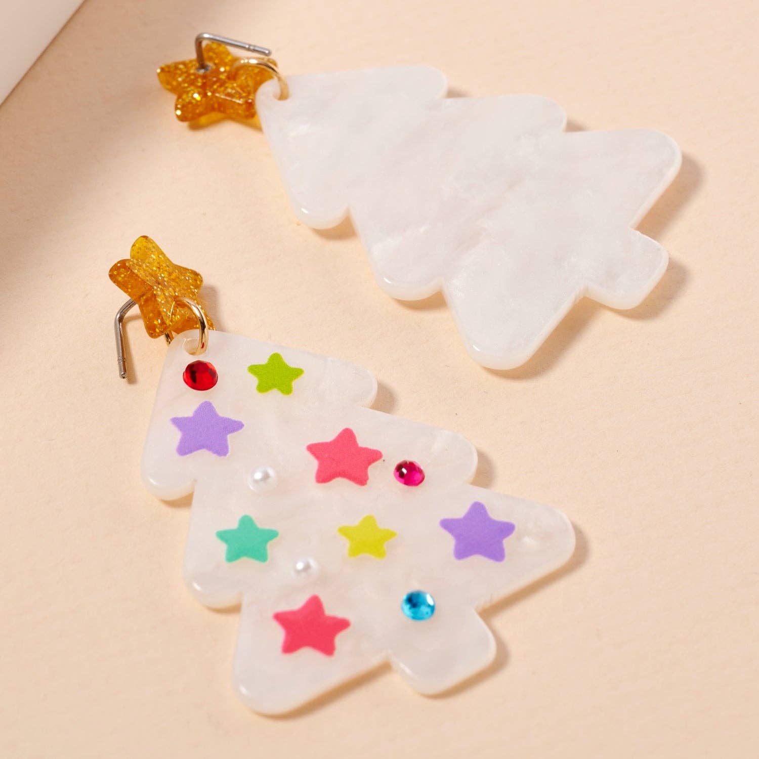 Add a festive touch to your holiday look with the Lilla Haven Glittery Christmas Tree Drop Earrings. This playful pair features two marbled white, acrylic tree-shaped earrings—one adorned with colorful stars and gems and the other left simple for contrast. Each is topped with a golden star, making these Holiday Acrylic Earrings a sparkling statement piece for any seasonal ensemble.