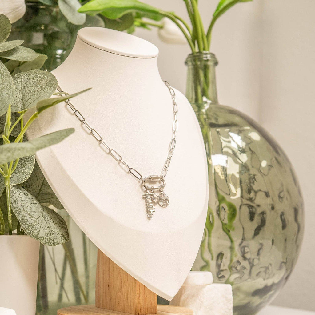 An 18K Gold PVD Paperclip Carabiner Necklace by WJW is displayed on a white bust, surrounded by green plants and a large glass vase in the background. This customizable necklace effortlessly combines elegance and versatility, making it ideal for personalizing your look.