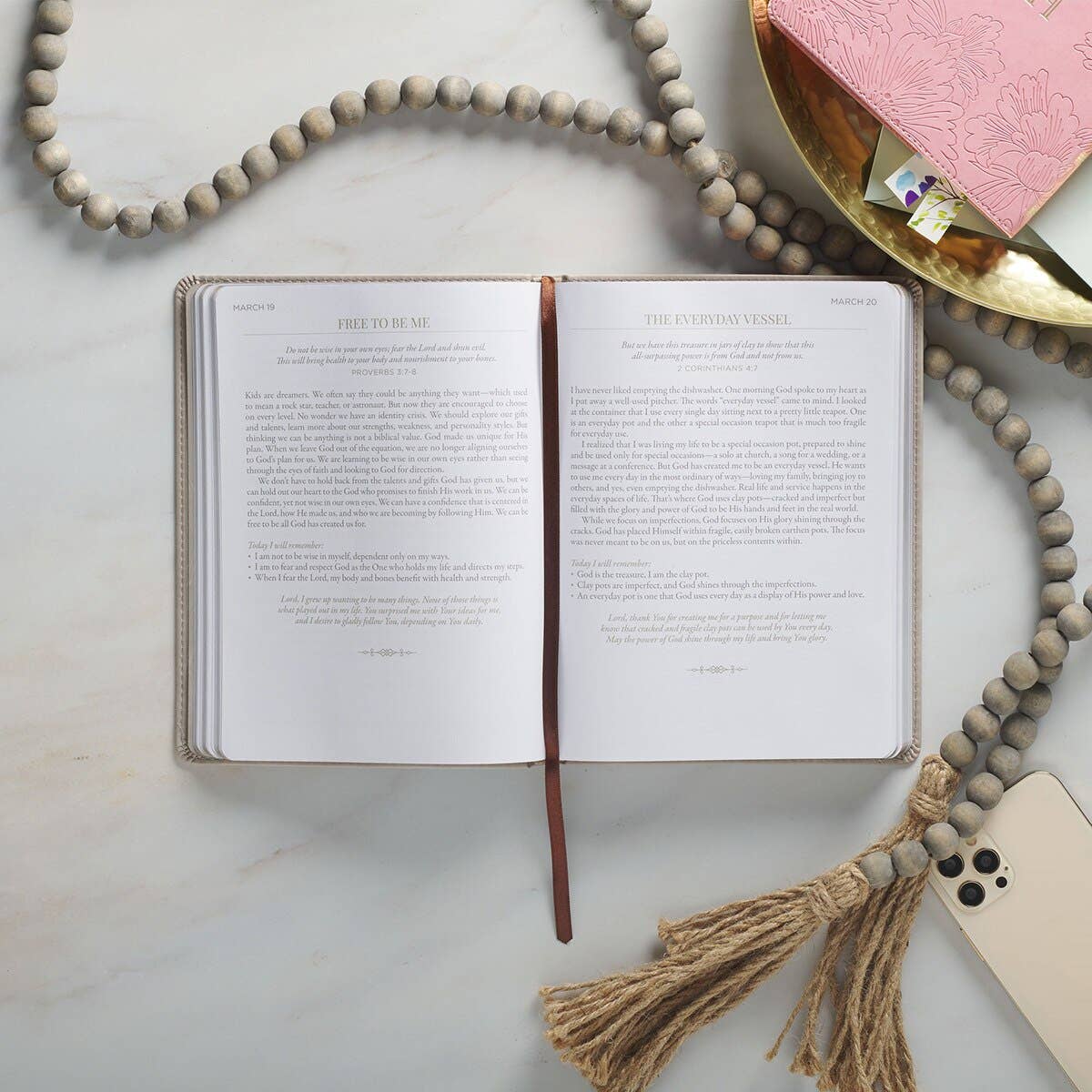 An open journal rests on a marble surface beside a beaded tassel, a gold tray holding a pink notebook, and a smartphone—setting the perfect scene for ten minutes of reflection with the Devotional 10 Minutes with God for Women Faux Leather by Christian Art Gifts during your daily quiet time.