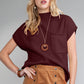 A woman with blonde hair wears a maroon Shewin Patch Pocket Ribbed Knit Short Sleeve Sweater, ideal for transitional weather, paired with a camel-colored skirt and a round pendant necklace. She stands against a gray background.