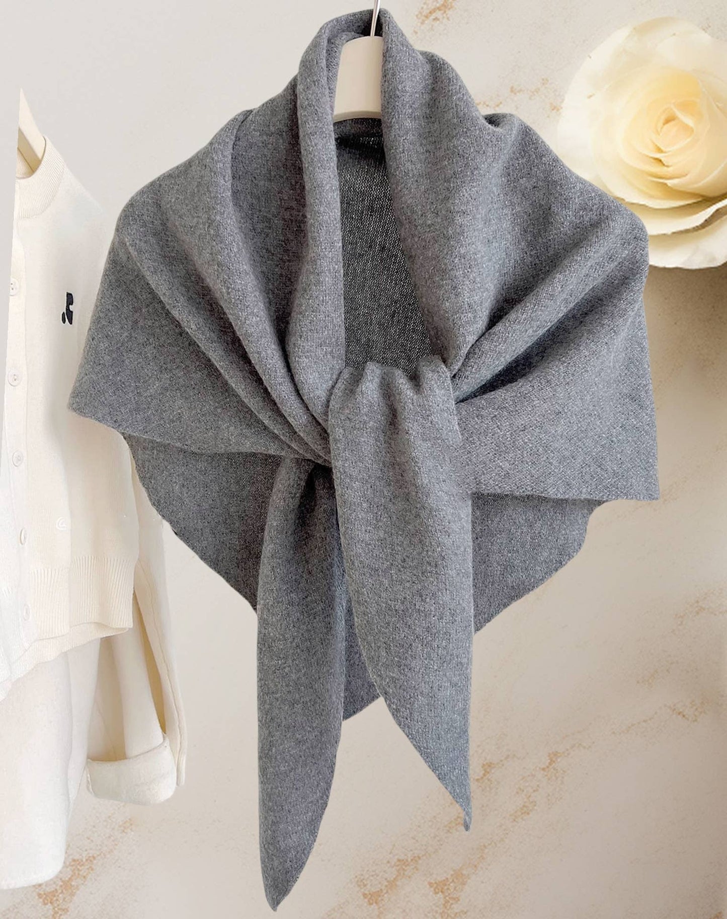 The JC Sunny Fashion Cashmere-Blend Knit Triangle Shawl with Cross-Front Design in gray is styled on a white hanger beside a cream sweater and large white faux flower, delivering chic elegance to any outfit.